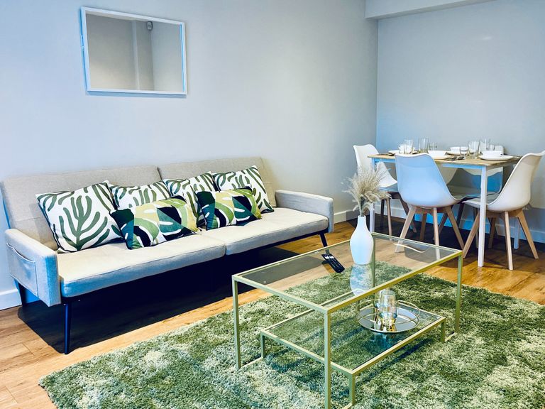 Modern living room, beige sofa and green foliage patterned cushions, fluffy olive rug, gold trimmed coffee table, wall mirror and full dining set. 